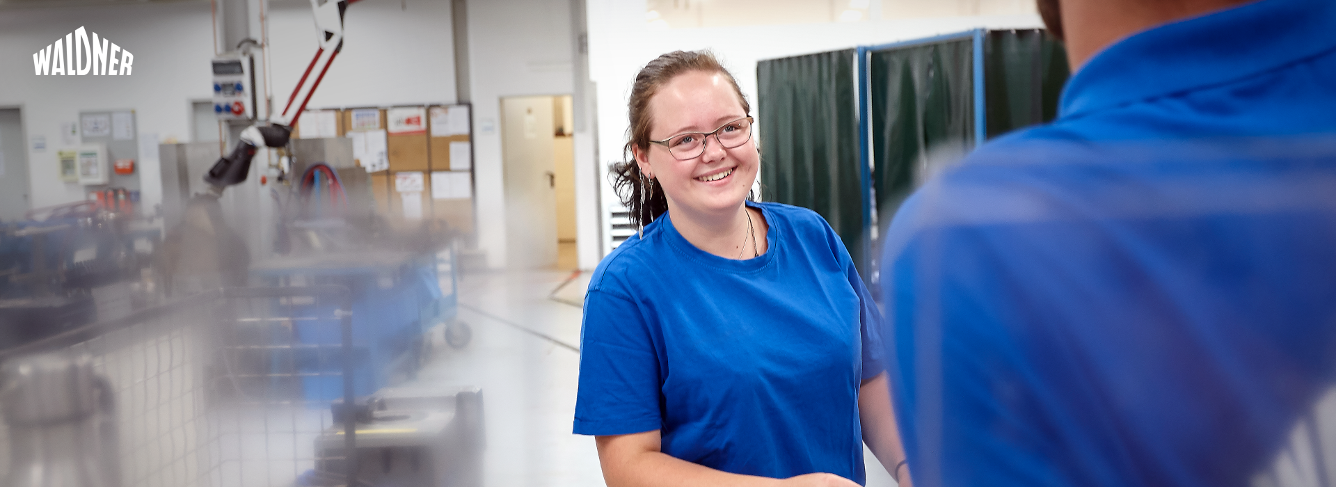 Im Bildzentrum steht eine Frau in einem blauen Shirt, die sich angeregt mit einem Kollegen unterhält.